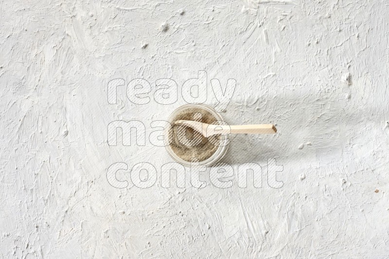 A glass jar and wooden spoon full of white pepper powder on textured white flooring