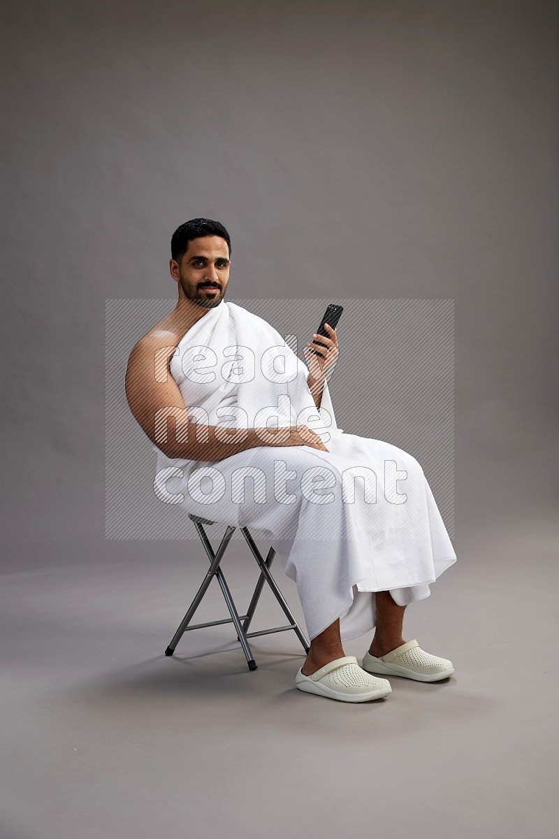 A man wearing Ehram sitting on chair texting on phone on gray background