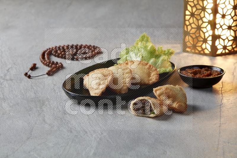 Three fried sambosas in an oval shaped black plate and a red sauce in a black round ramekin with a brown misbaha and a golden lantern on the side on a gray background