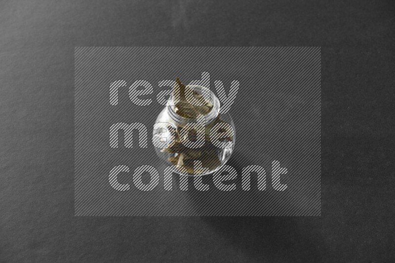 A glass spice jar full of bay laurel leaves on black flooring