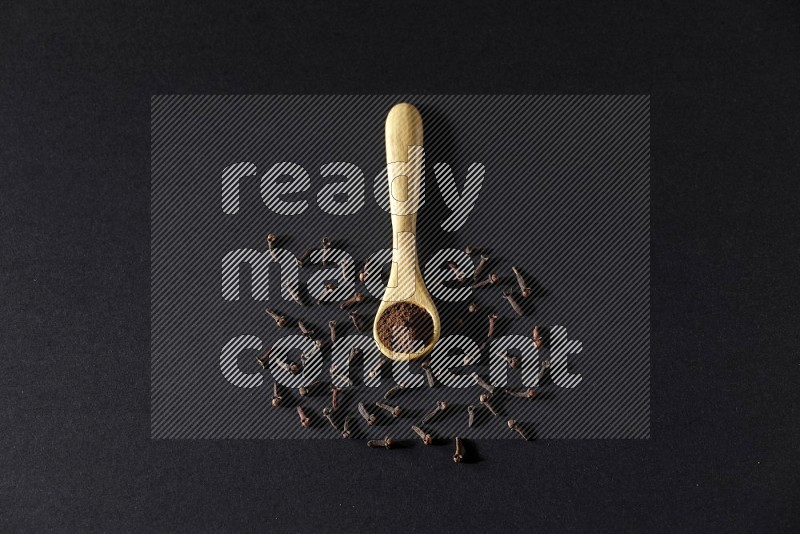 A wooden spoon full of cloves powder with spreaded whole cloves on a black flooring