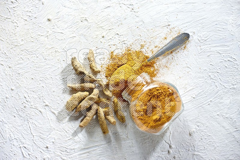 A flipped glass spice jar and metal spoon full of turmeric powder and powder spilled out of it with dried whole fingers on textured white flooring