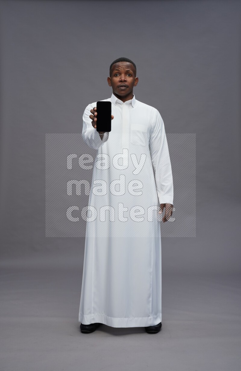 Saudi man wearing thob standing showing phone to camera on gray background