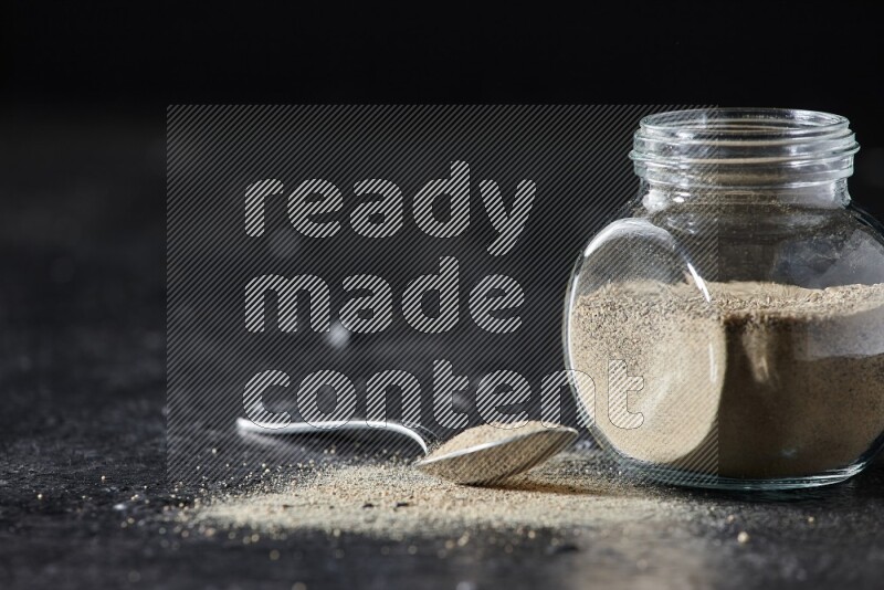Herbal glass jar and metal spoon full of white pepper powder on textured black flooring