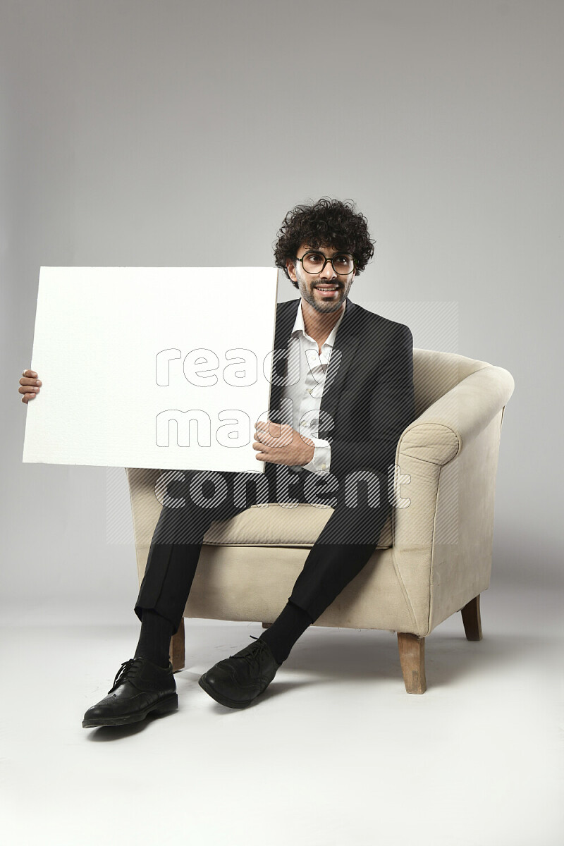 رجل يرتدي ملابس رسمية يجلس علي كرسي و يحمل لوحة بيضاء علي خلفية بيضاء