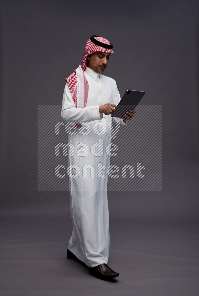 Saudi man wearing thob and shomag standing working on tablet on gray background