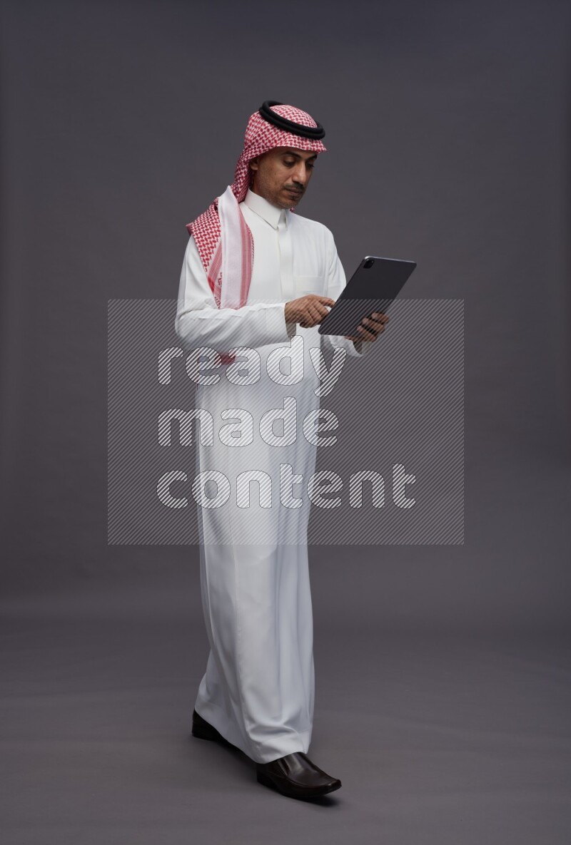 Saudi man wearing thob and shomag standing working on tablet on gray background
