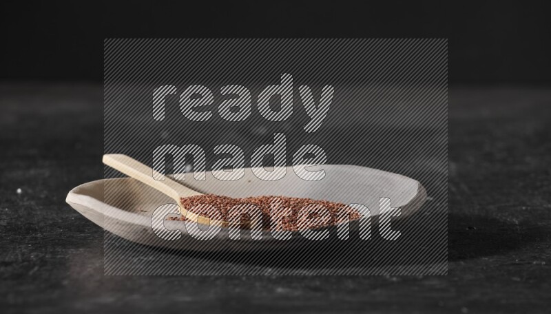 A multicolored pottery plate full of garden cress seeds and wooden spoon full of seeds on a textured black flooring