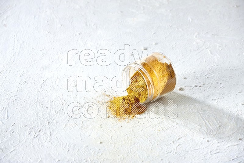 A flipped glass spice jar full of turmeric powder and powder fell out it on textured white flooring