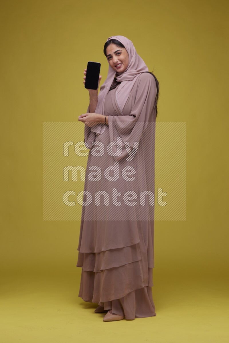 A woman Showing her Phone screen on a Yellow Background wearing Brown Abaya with Hijab