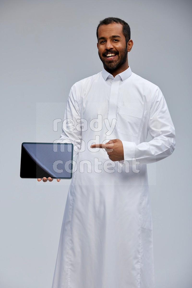 Saudi man wearing Thob standing showing tablet to camera on Gray background