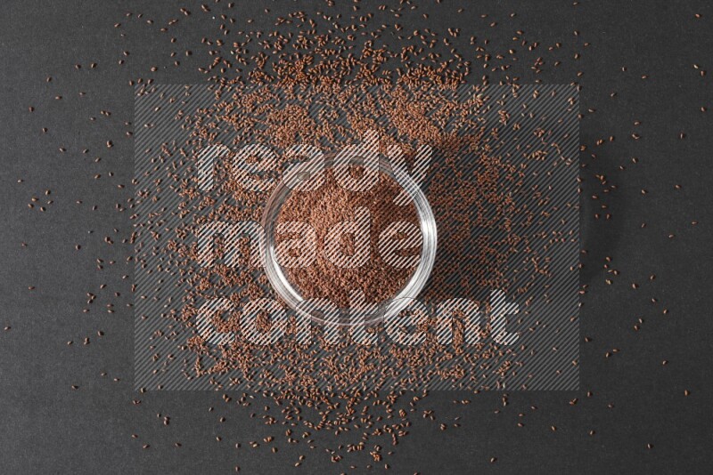 A glass bowl full of garden cress seeds surrounded by seeds on a black flooring