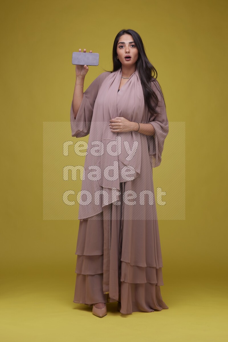 A woman Showing SmartPhone Screen on a Yellow Background wearing Brown Abaya