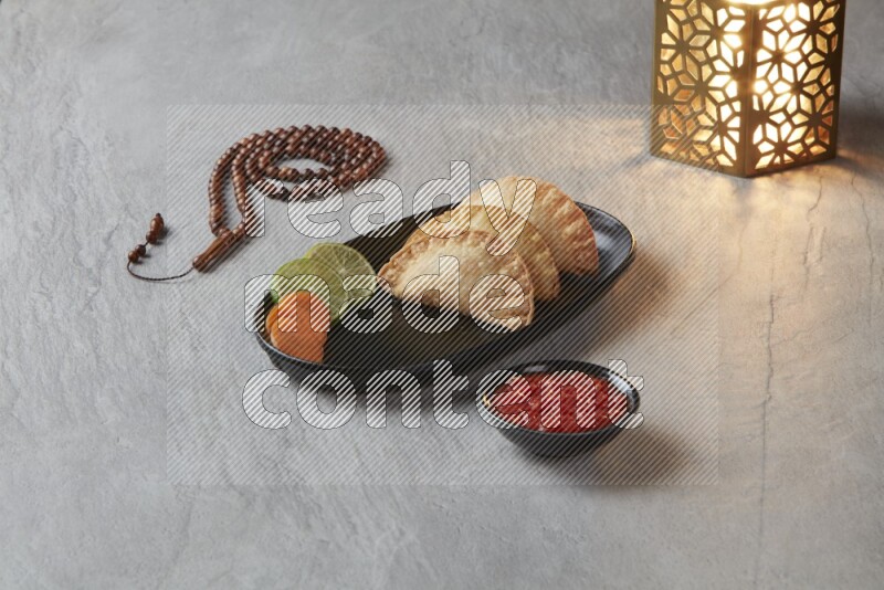 Three fried sambosas in an oval shaped black plate and a red sauce in a black round ramekin with a brown misbaha and a golden lantern on the side on a gray background