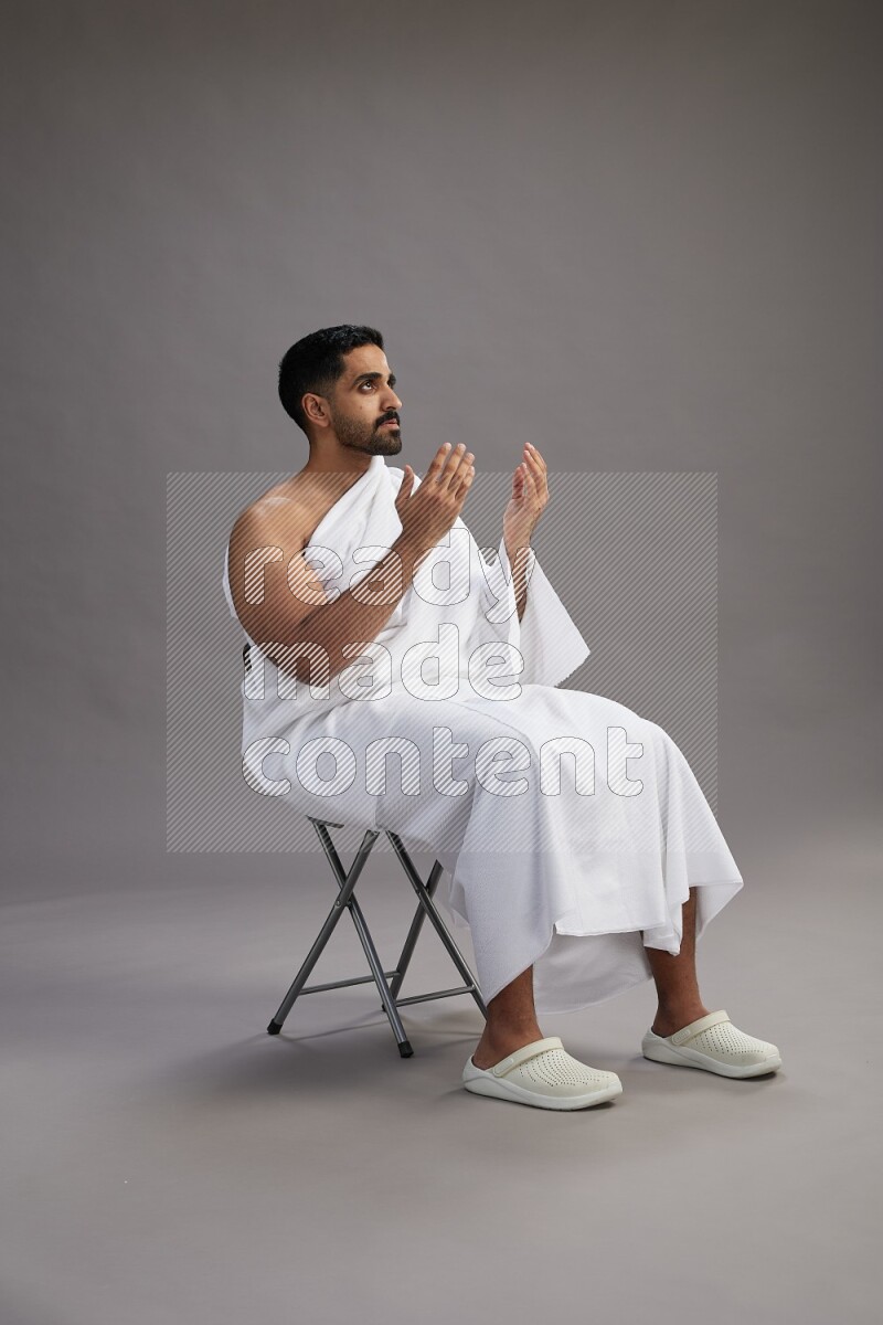 A man wearing Ehram sitting on chair performing dua'a on gray background