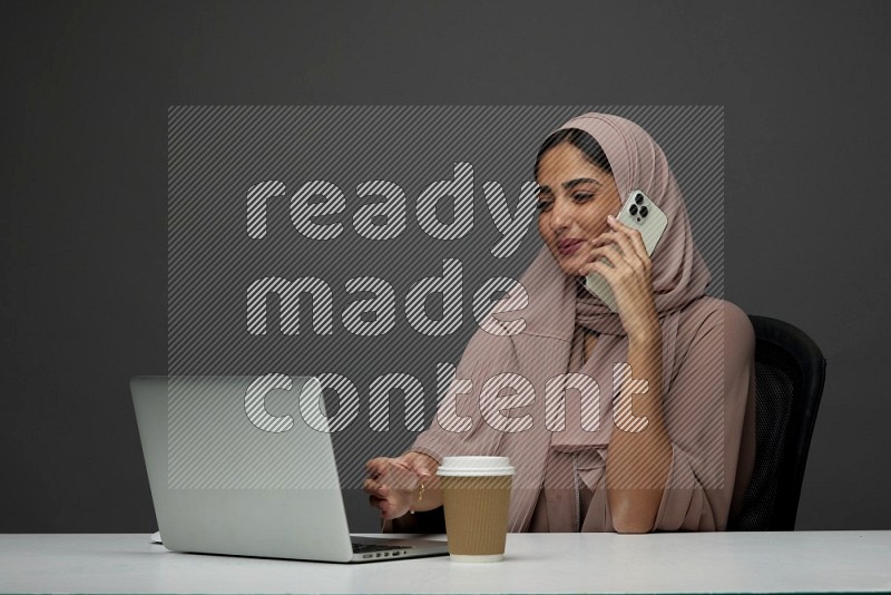 A Saudi woman Setting on her desk
 calling  on a Gray Background wearing Brown Abaya with Hijab