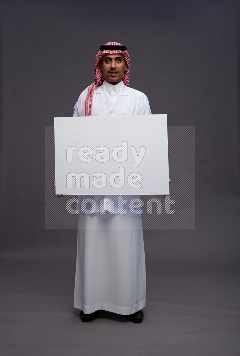 Saudi man wearing thob with lab coat and shomag with pocket employee badge standing holding board on gray background