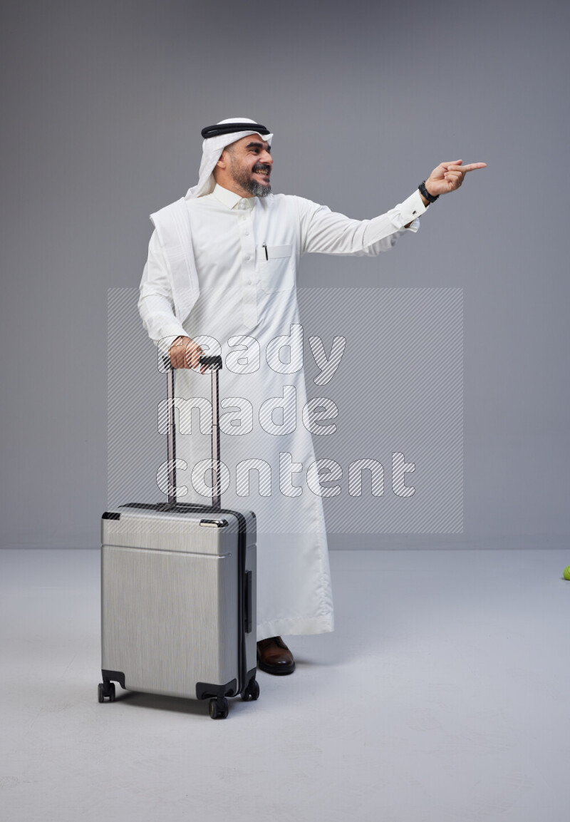 Saudi man wearing Thob and white Shomag standing holding Travel bag on Gray background