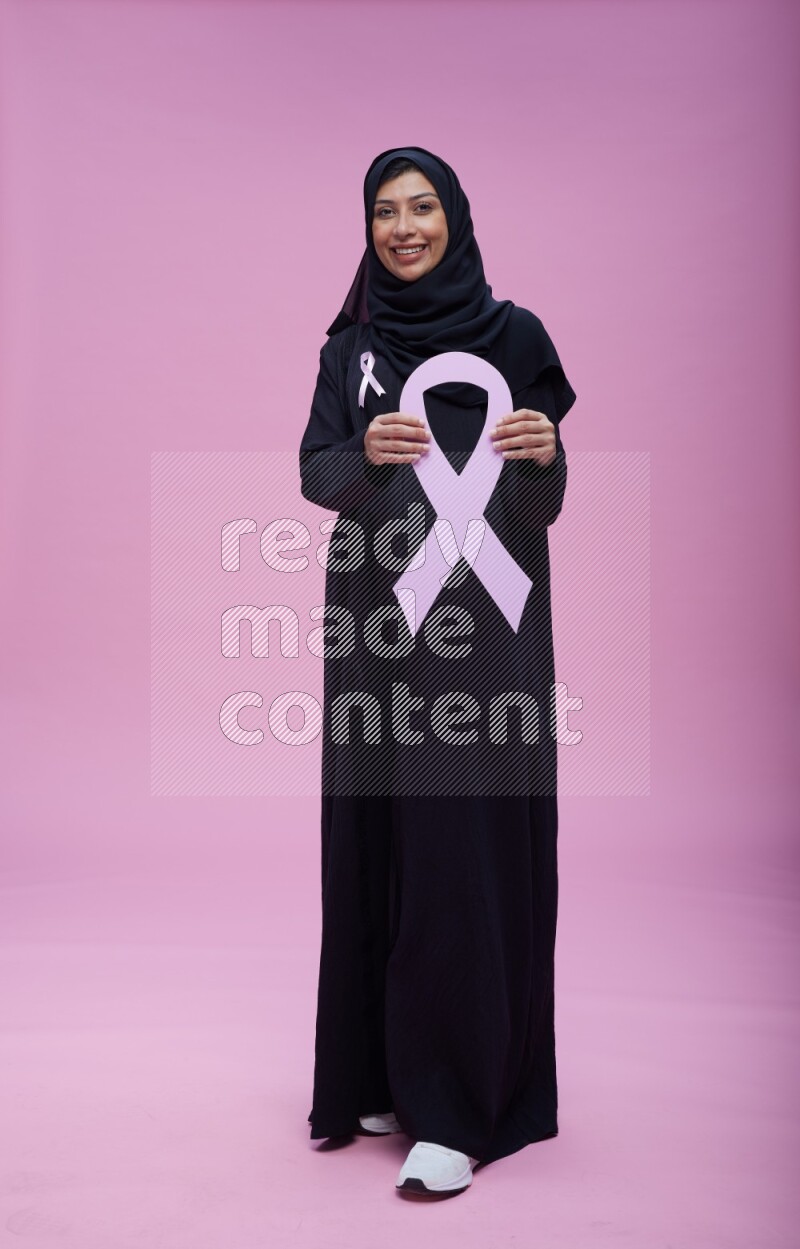 Saudi woman wearing pink ribbon on abaya standing holding awareness ribbon on plain pink background