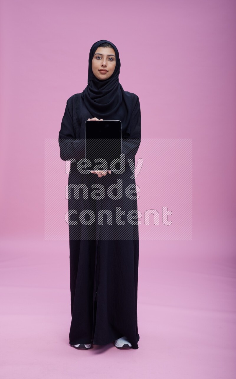 Saudi woman wearing Abaya standing showing tablet to camera on pink background