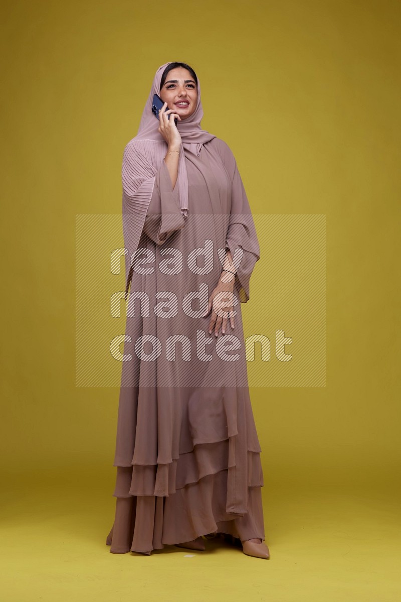 A woman having a call on a Yellow Background wearing Brown Abaya with Hijab