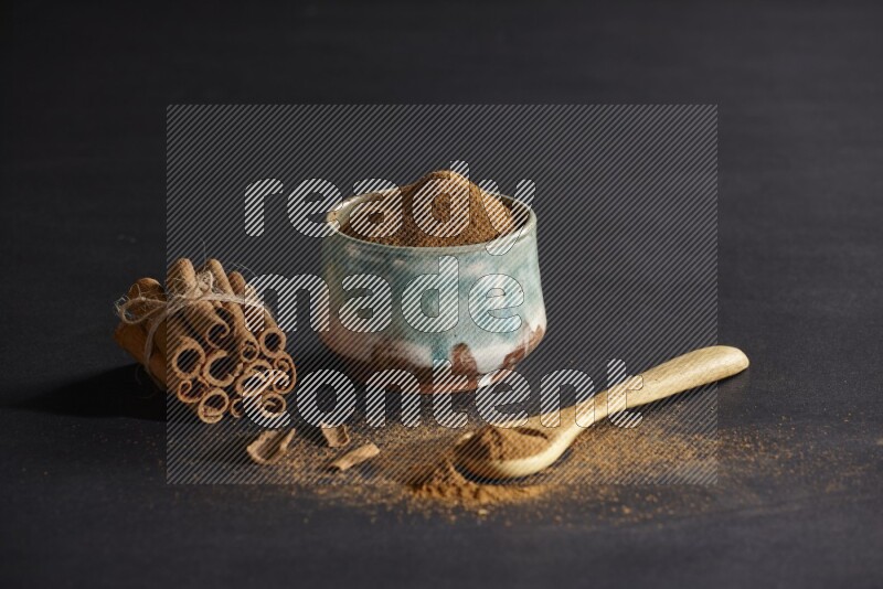 Ceramic bowl full of cinnamon powder and a wooden spoon full of powder with cinnamon sticks stacked and bounded on black background