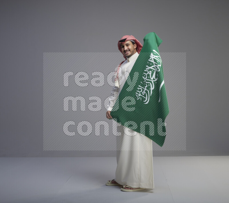 A saudi man standing wearing thob and red shomag holding big saudi flag on gray background
