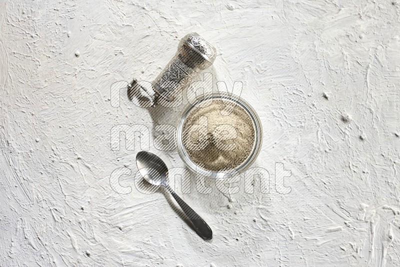 وعاء زجاجي ممتلئ ببودرة الفلفل الأبيض مع حبوب الفلفل الأبيض ومطحنة معدنية على أرضية بيضاء