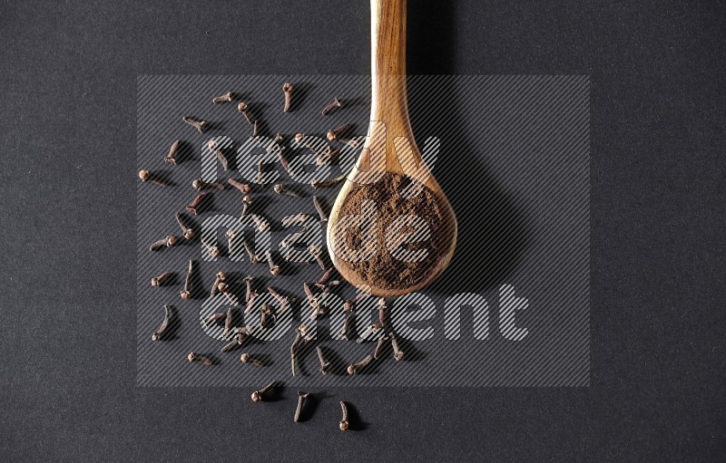 A wooden ladle full of cloves powder and some of whole cloves around it on a black flooring