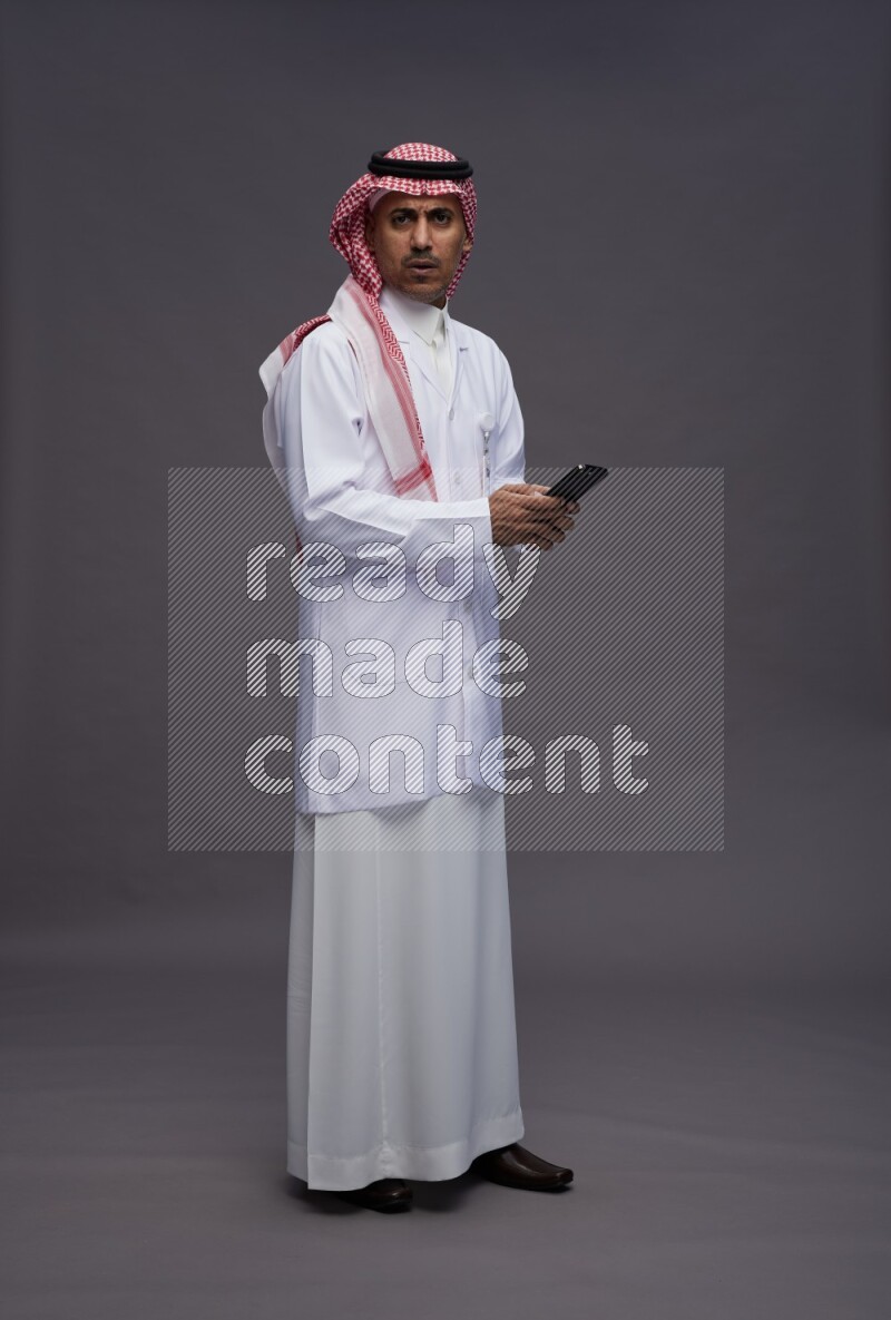 Saudi man wearing thob with lab coat and shomag with pocket employee badge standing texting on phone on gray background