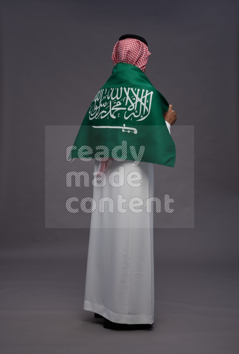 Saudi man wearing thob and shomag standing holding Saudi flag on gray background