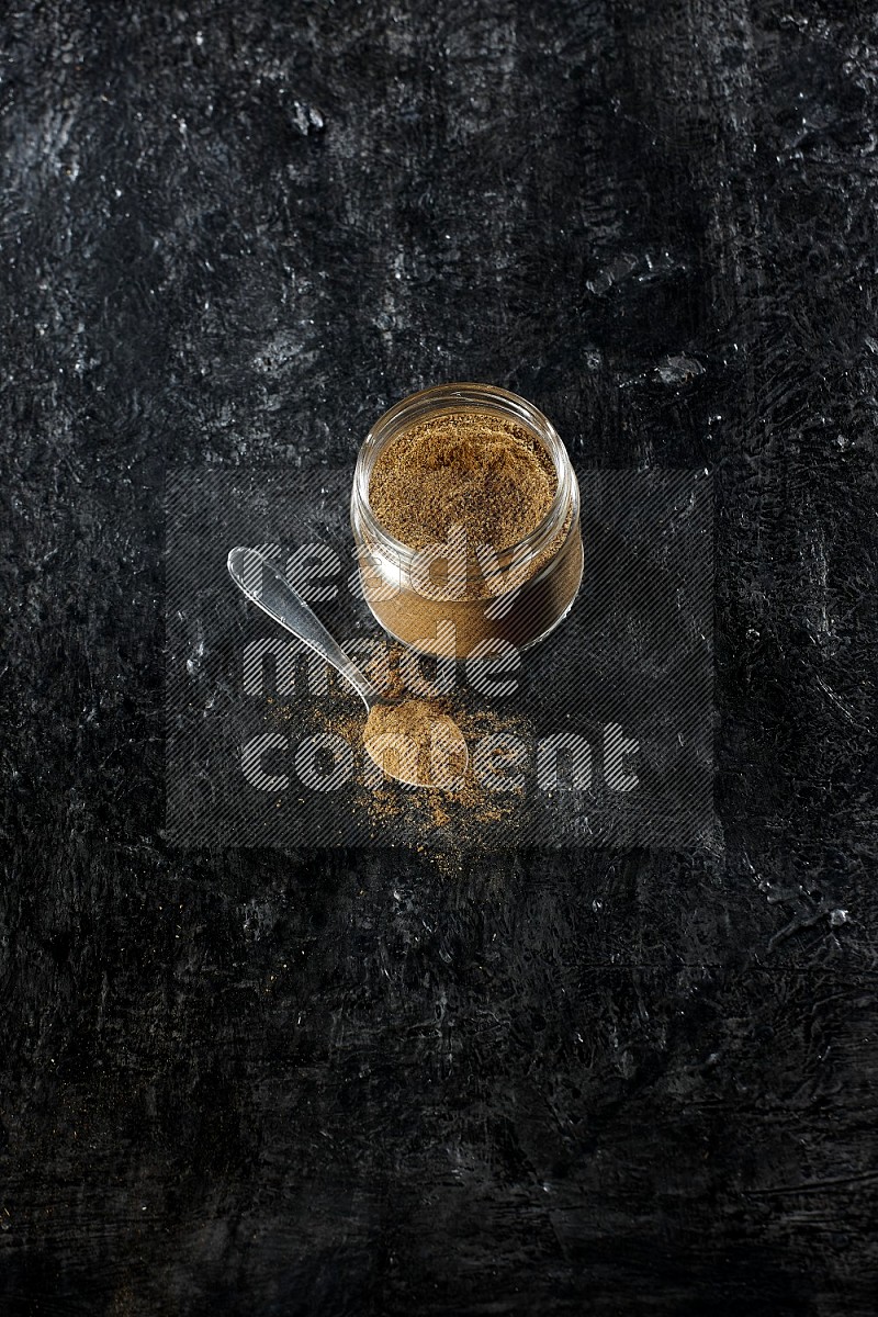 A glass jar and a metal spoon full of cumin powder on a textured black flooring