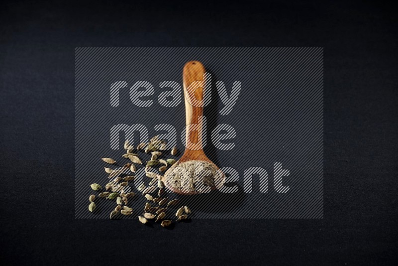 A wooden ladle full of cardamom powder and cardamom seeds beside it on black flooring