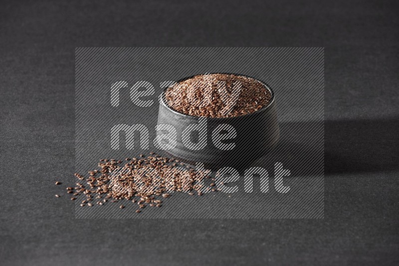 A black pottery bowl full of flaxseeds surrounded by the seeds on a black flooring