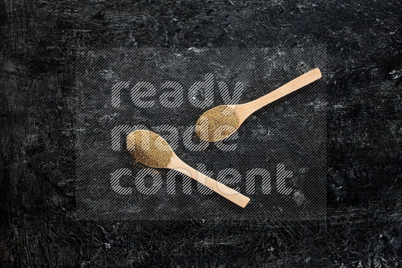 2 wooden spoons full of cumin powder on textured black flooring
