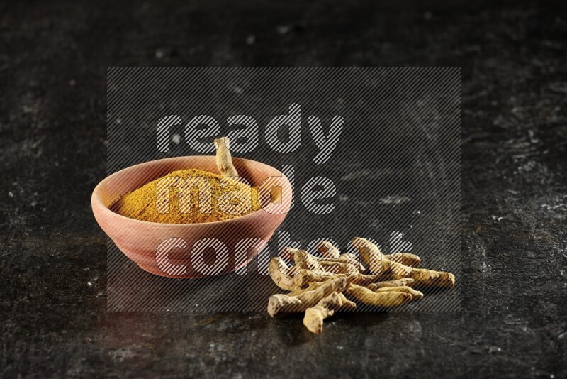 A wooden bowl full of turmeric powder with dried turmeric whole fingers on textured black flooring