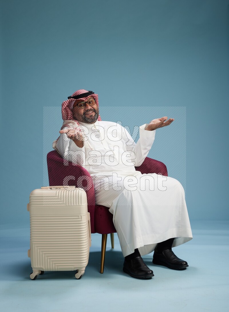 Saudi Man with shimag sitting on chair with a travel luggage on blue background