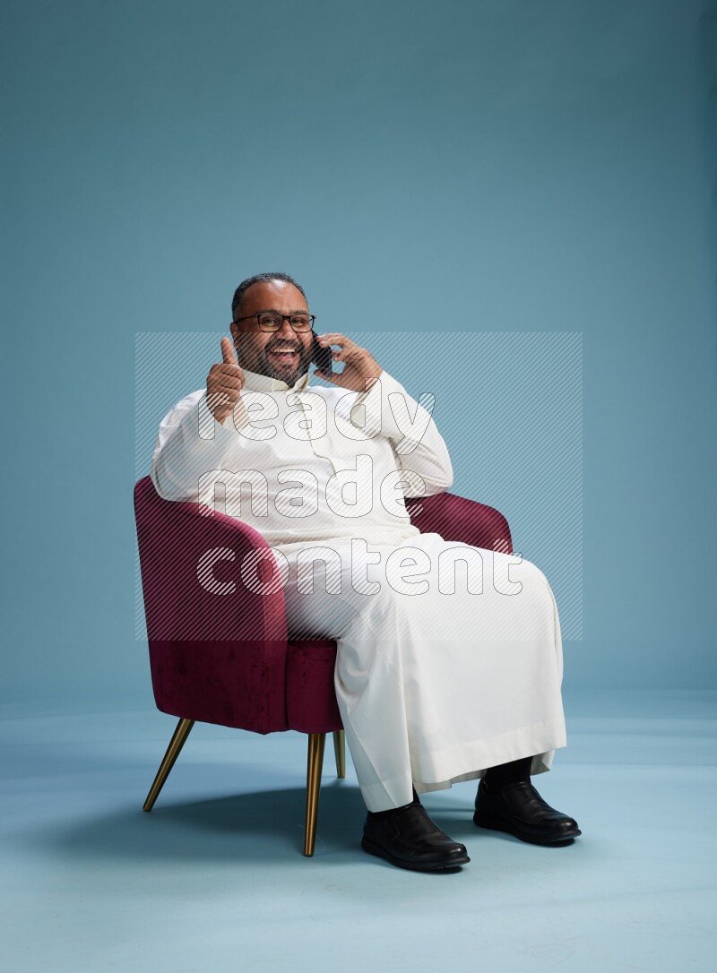 Saudi Man without shimag sitting on chair talking on phone on blue background
