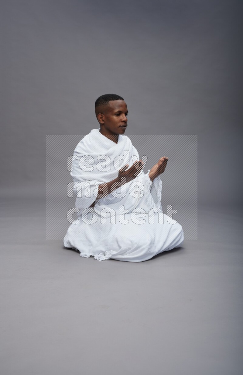 A man wearing Ehram sitting on floor dua'a on gray background