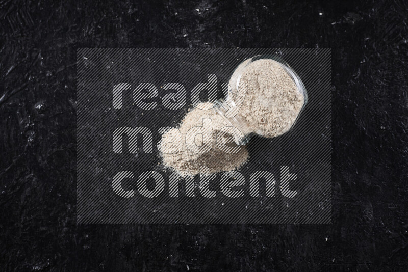 A glass jar full of onion powder flipped with some spilling powder on black background