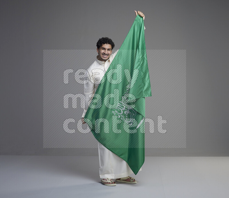 A Saudi man standing wearing thob holding big Saudi flag on gray background