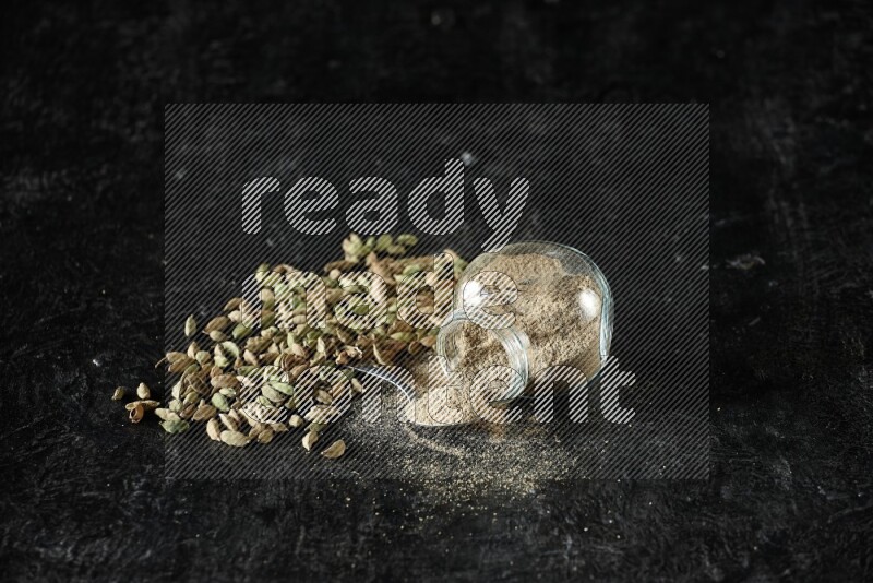 A flipped glass spice jar and a metal spoon full of cardamom powder and cardamom seeds spreaded on textured black flooring