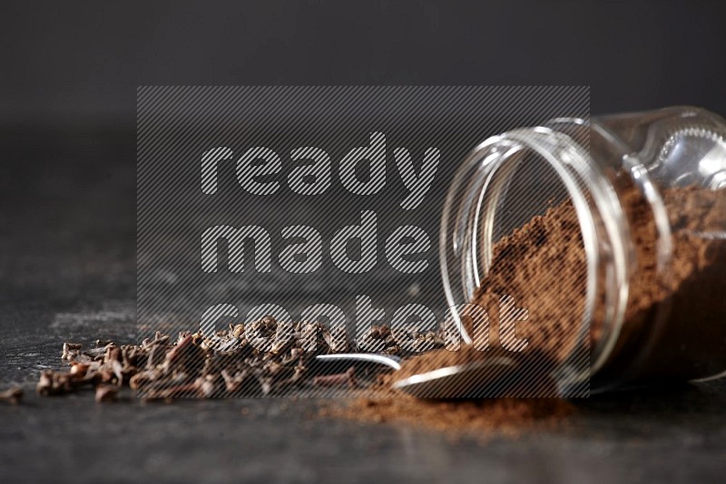 A flipped glass jar and metal spoon full of cloves powder with cloves spread on a textured black flooring