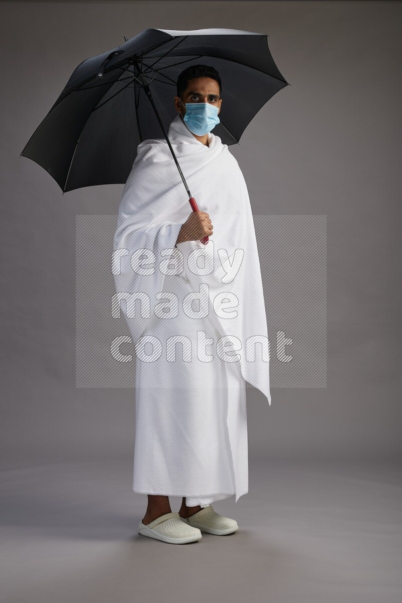 A man wearing Ehram and mask Standing holding umbrella on gray background on gray background