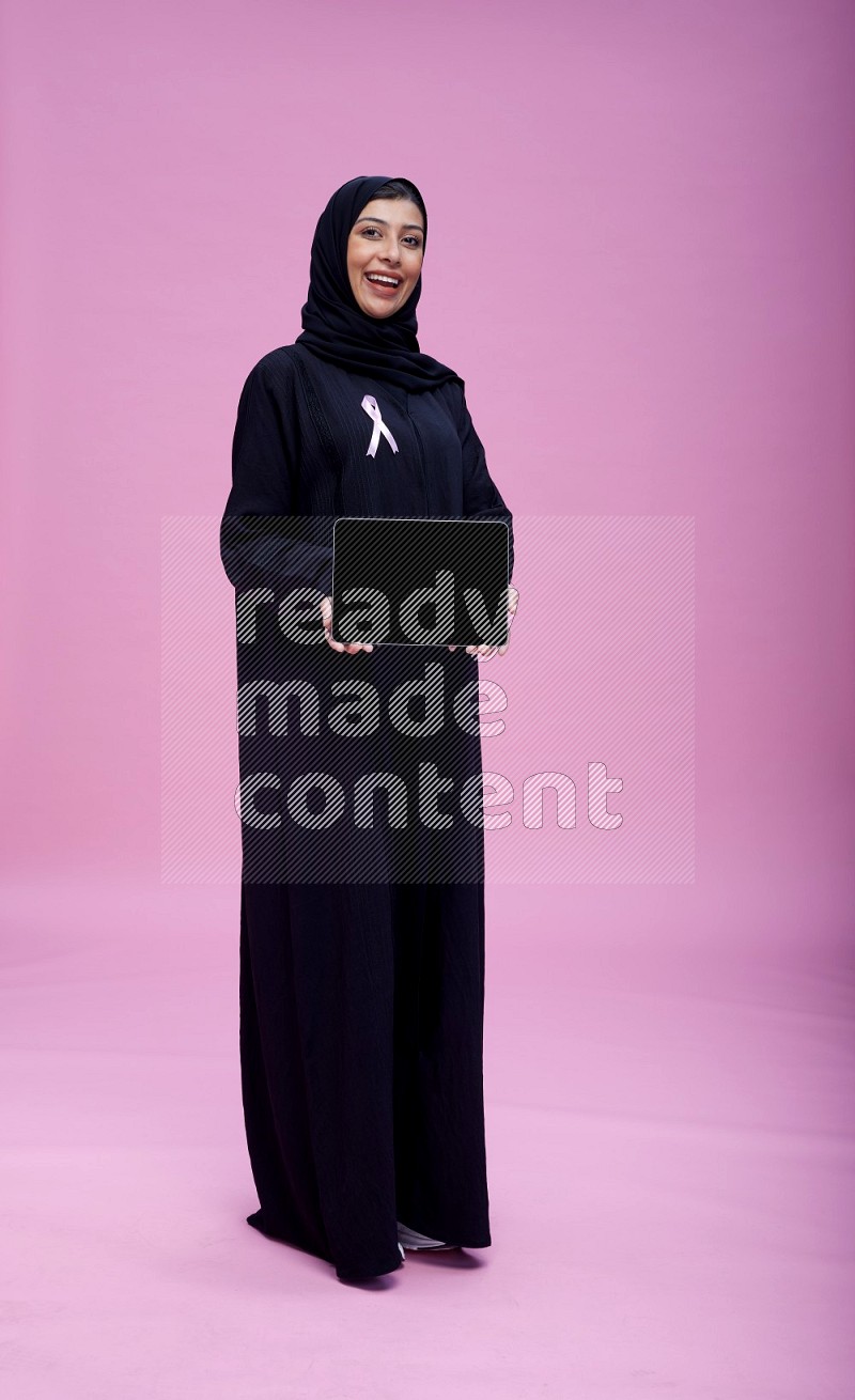 Saudi woman wearing pink ribbon on Abaya standing showing tablet to camera on pink background