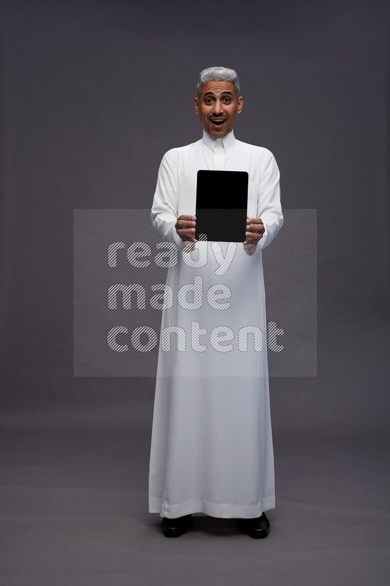 Saudi man wearing thob with neck strap employee badge standing showing tablet to camera on gray background