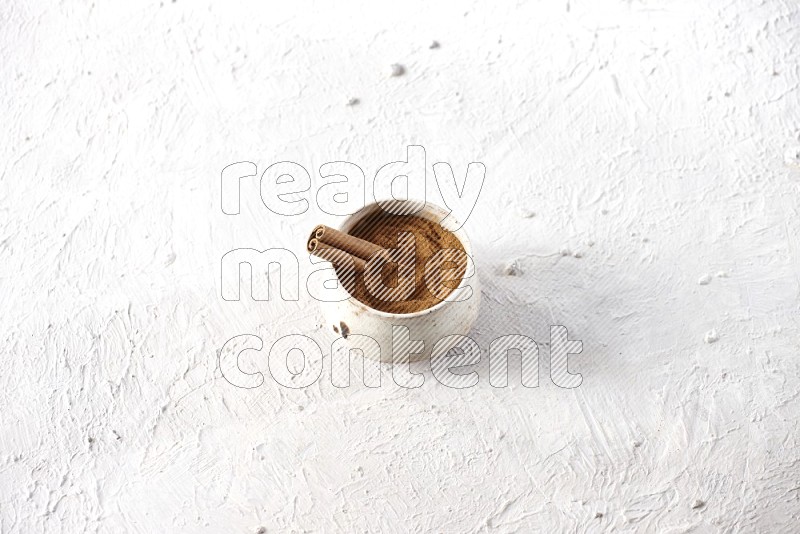 Ceramic beige bowl full of cinnamon powder with a cinnamon stick on a textured white background