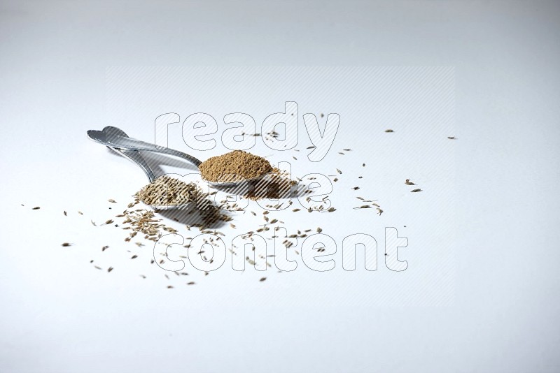 2 metal spoons full of cumin seeds and powder on white flooring