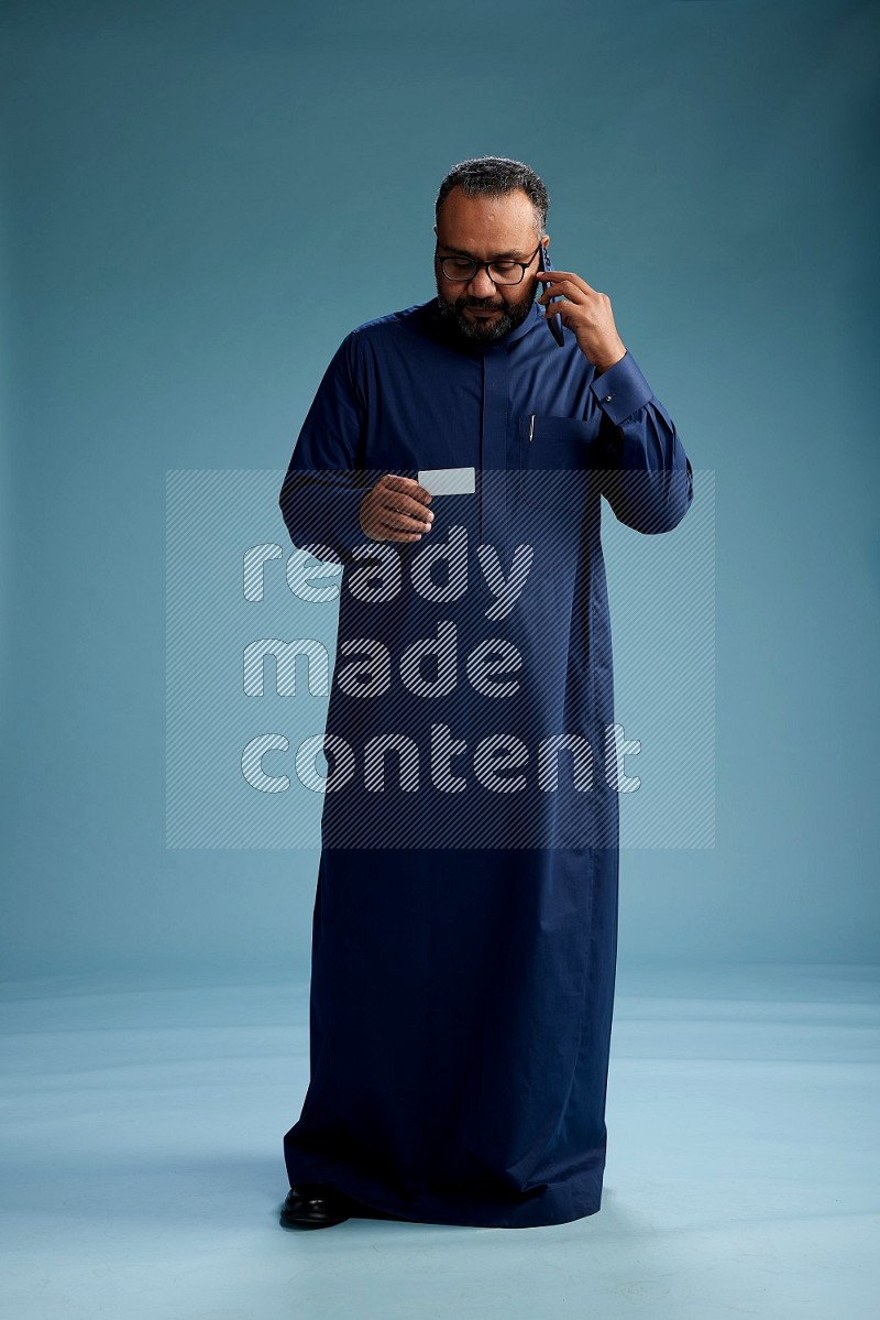 Saudi Man without shimag Standing holding ATM while talking on phone on blue background