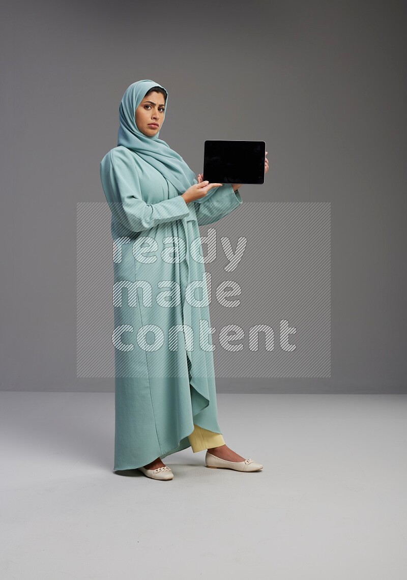 Saudi Woman wearing Abaya standing showing tablet to camera on Gray background